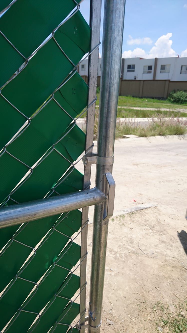 Primer plano de una puerta de alambre con listones verdes para privacidad y un poste de metal en un entorno al aire libre.
