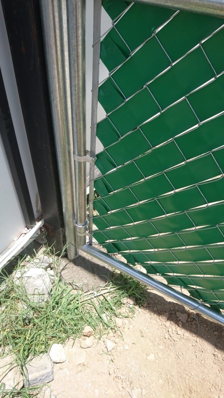 Valla metálica con lamas verdes junto a un edificio, con césped y tierra en su base.