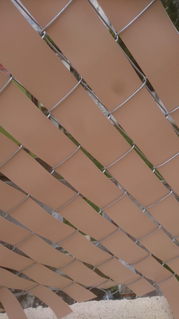 Láminas de privacidad de color tostado tejidas a través de una cerca de alambre, creando un patrón diagonal; el cielo es parcialmente visible.