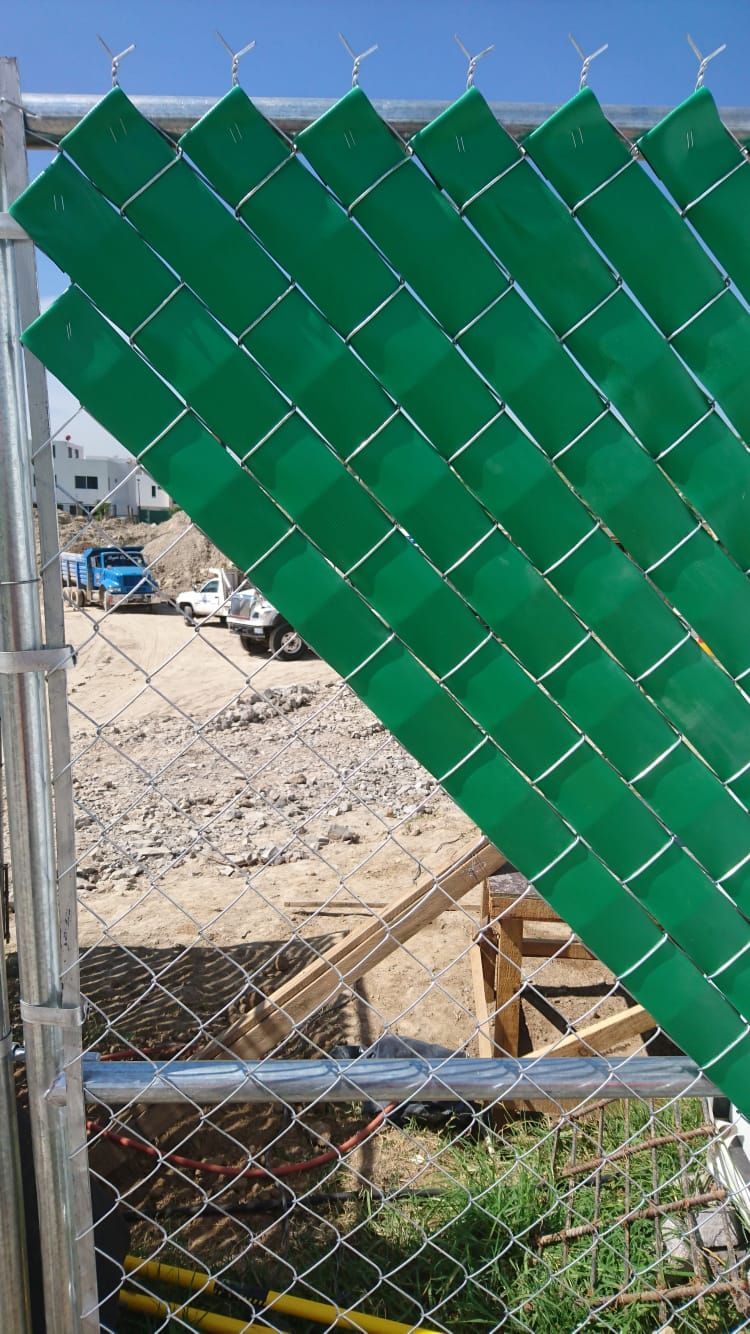 Listones verdes colocados en diagonal en una cerca de alambre. Obras en construcción al fondo.