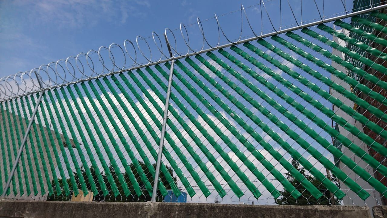 Valla verde con listones diagonales, rematada con alambre de púas, contra un cielo azul.