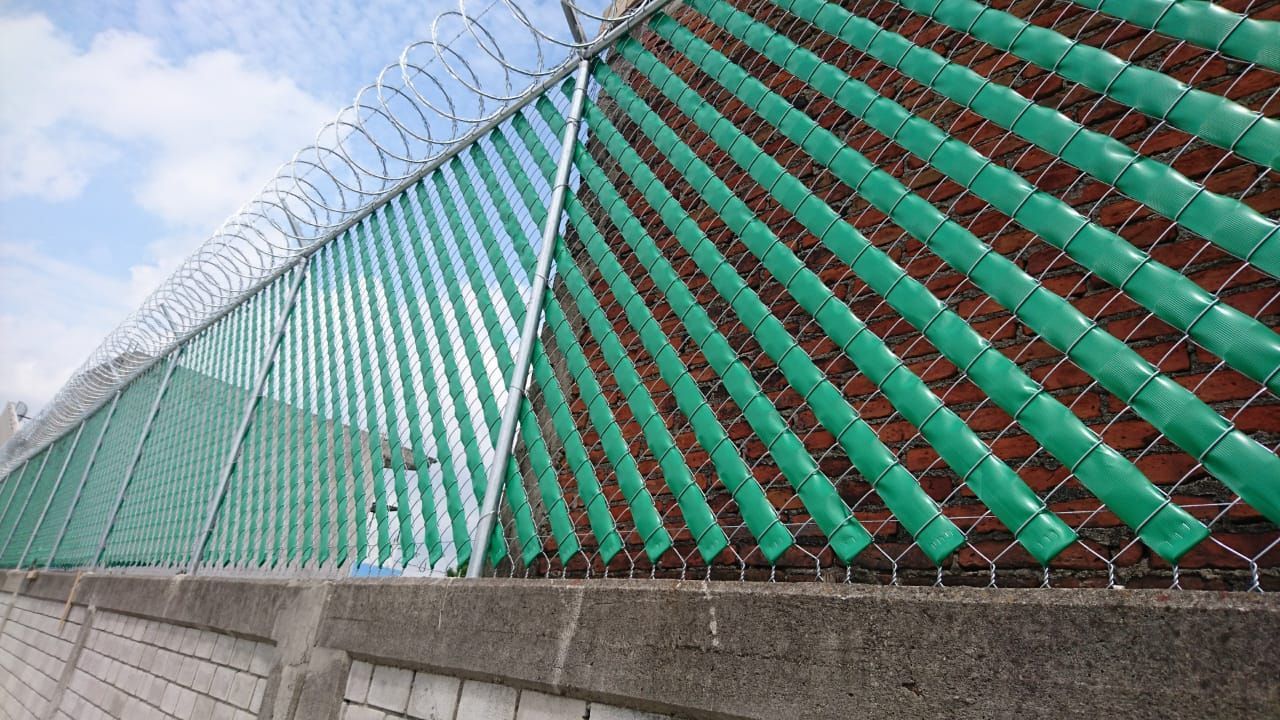 Valla de listones verdes con alambre de púas en la parte superior, contra una pared de ladrillos, al aire libre.