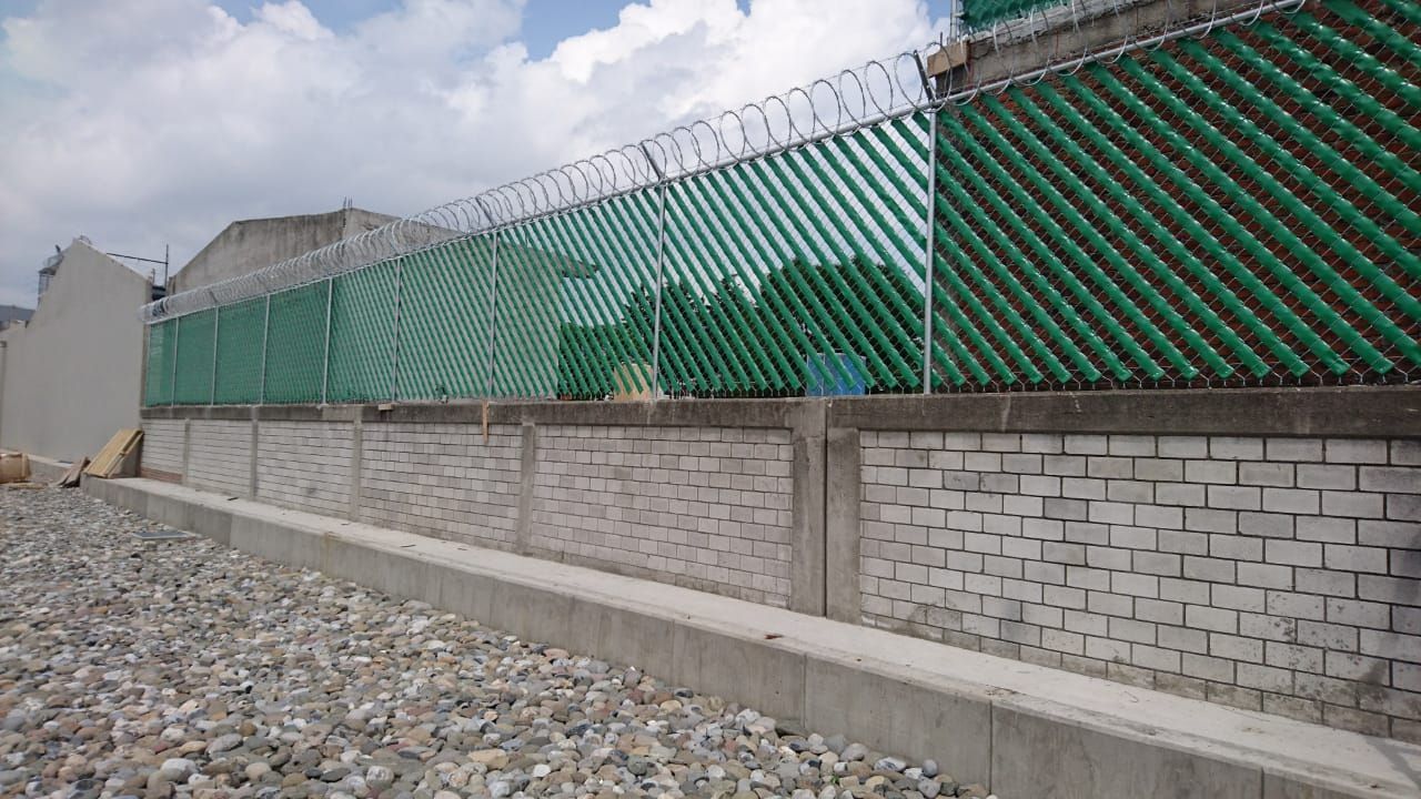 Una valla verde larga con alambre de púas encima, construida con botellas verdes reutilizadas.