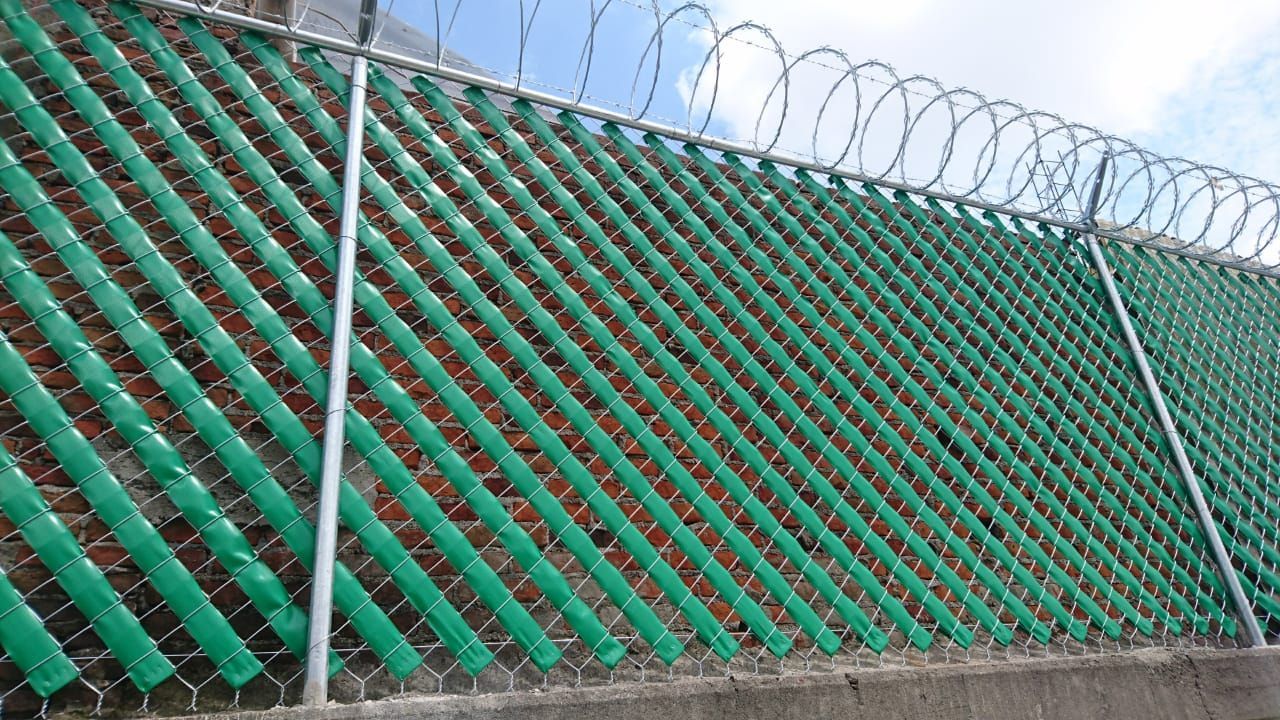 Listones verdes para privacidad tejidos en una cerca de alambre de cadena rematada con alambre de púas, contra un cielo azul.