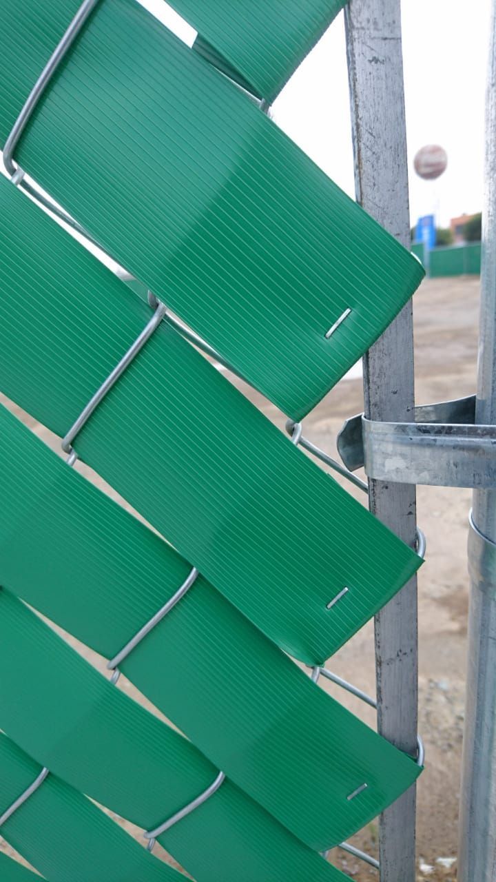 Listones de plástico verde tejidos en una valla de alambre junto a un poste de metal.