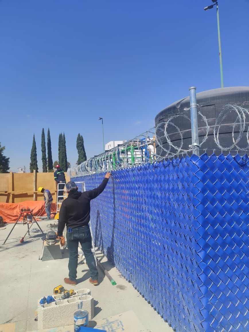 Trabajadores instalando una barrera de plástico azul junto a un tanque de agua, coronado con alambre de púas, en un día soleado.