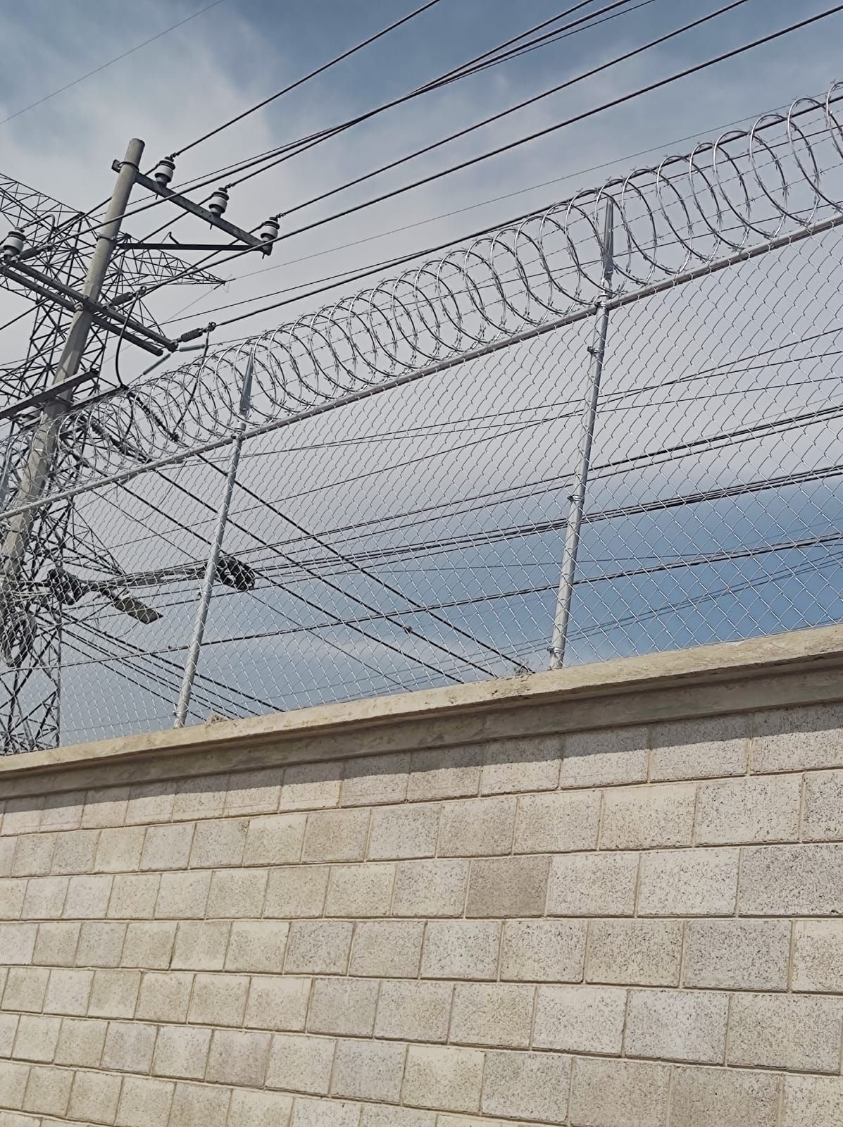 Valla de seguridad con alambre de púas sobre una pared de ladrillos, líneas eléctricas al fondo.