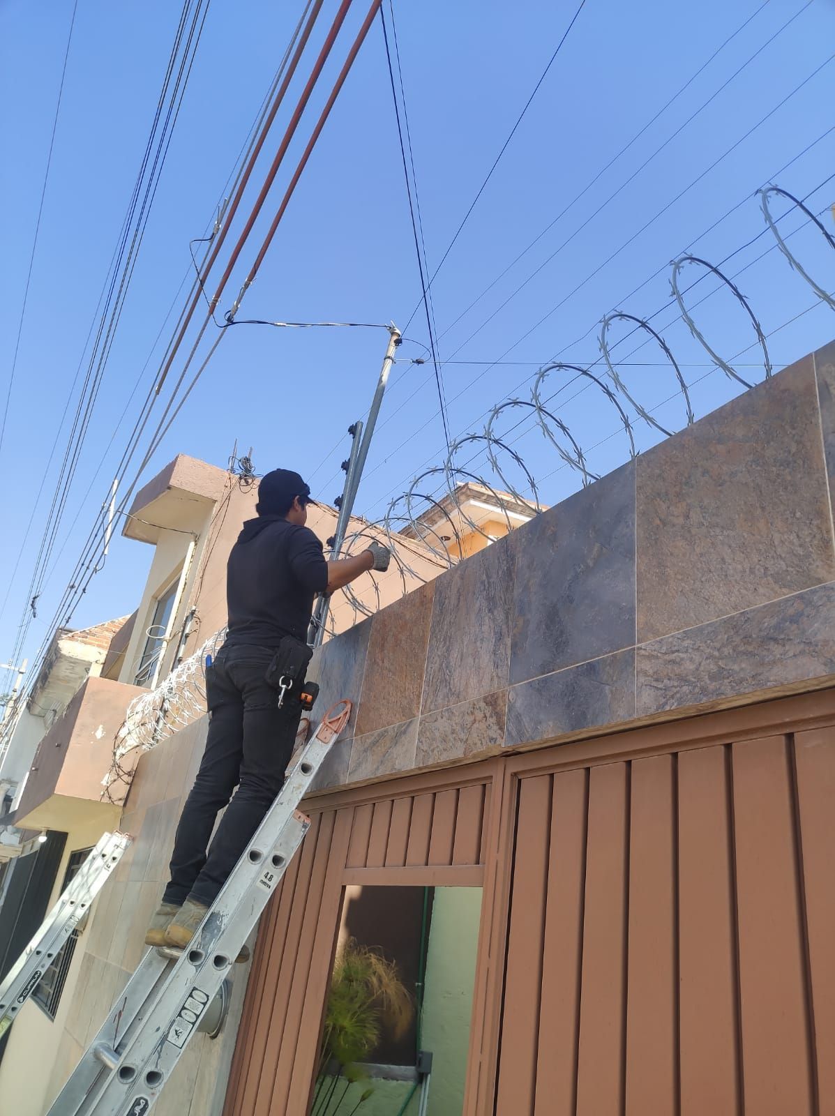 Hombre en una escalera instalando una valla de seguridad con alambre de púas en una pared.