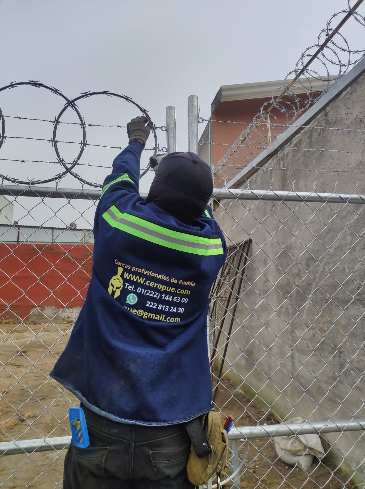 Hombre instalando alambre de púas en una cerca de alambre de cadena, vestido con una chaqueta de trabajo azul, al aire libre cerca de un muro de hormigón.