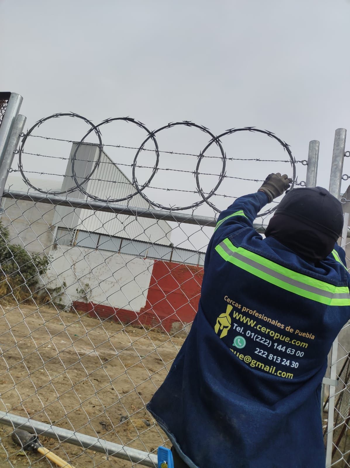 Trabajador instalando alambre de púas en una cerca de alambre de cadena; viste ropa de trabajo azul con rayas verdes y un gorro negro.