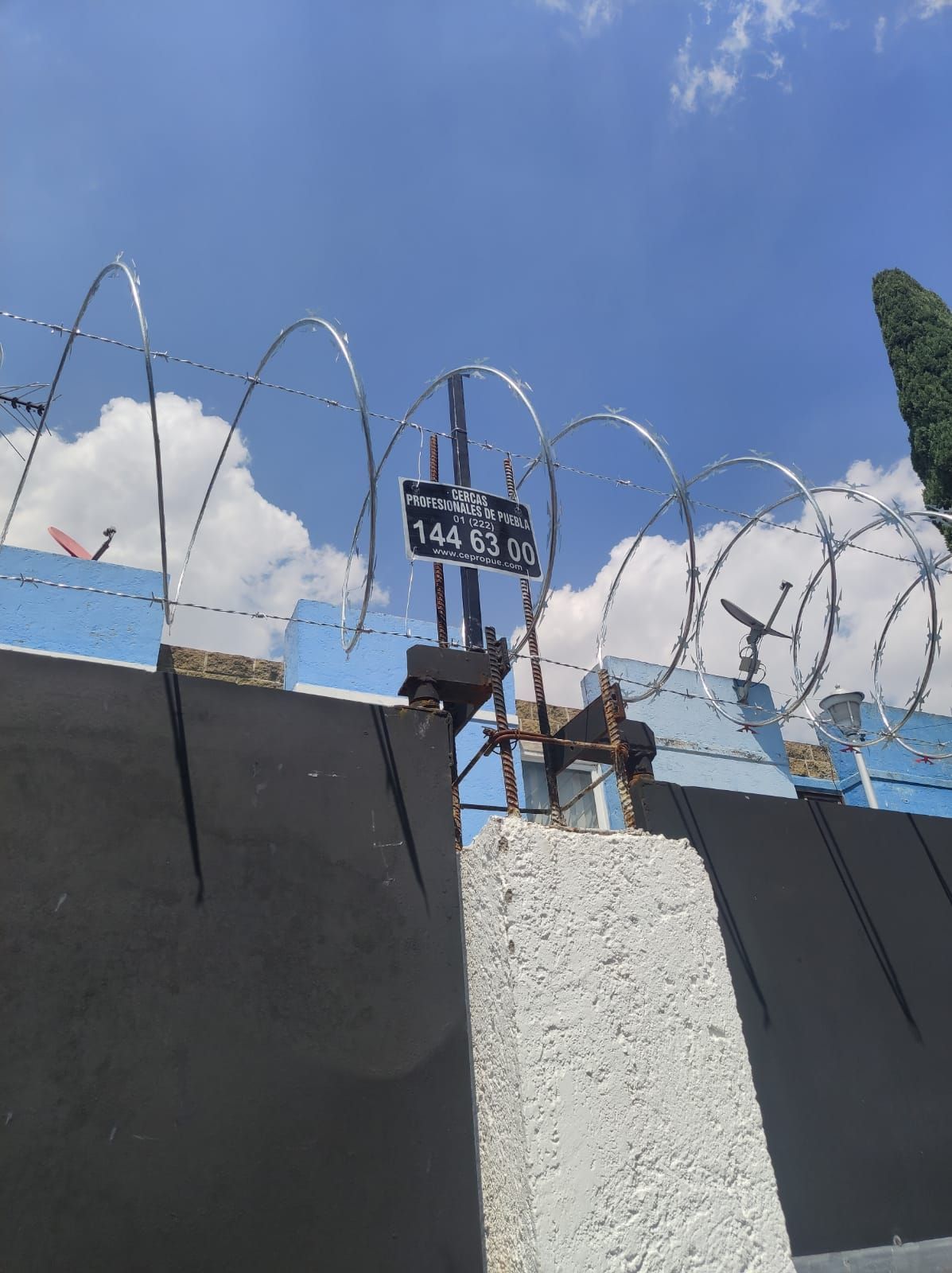 Una valla de hormigón coronada con alambre de púas bajo un cielo azul con un cartel adherido.