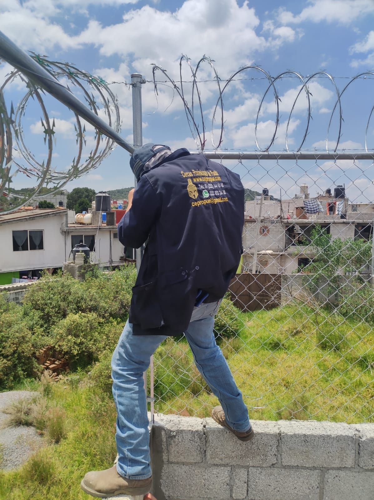 Hombre en un muro, alcanzando una barra de metal sobre una cerca de alambre de espino; camisa azul y vaqueros. Al aire libre, en un entorno soleado.