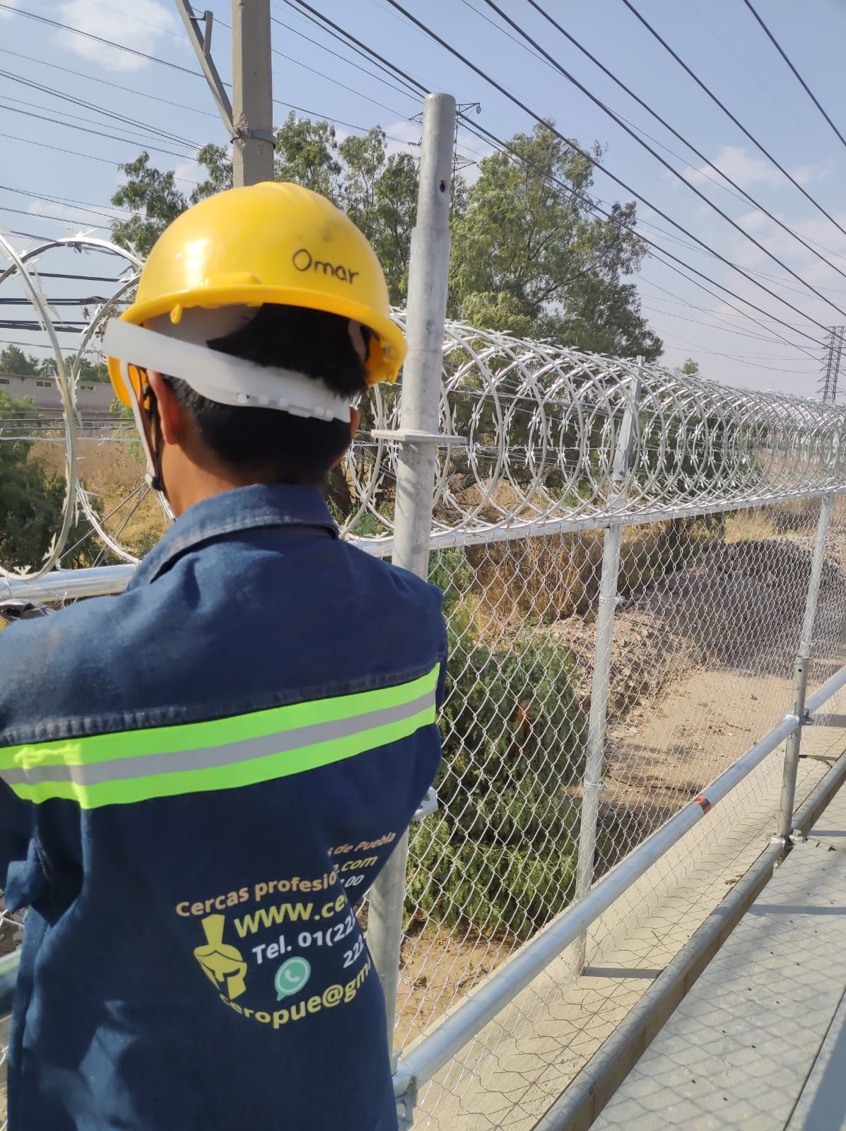Persona con uniforme de trabajo inspeccionando una cerca de alambre de púas. Uniforme azul y casco amarillo, al aire libre.