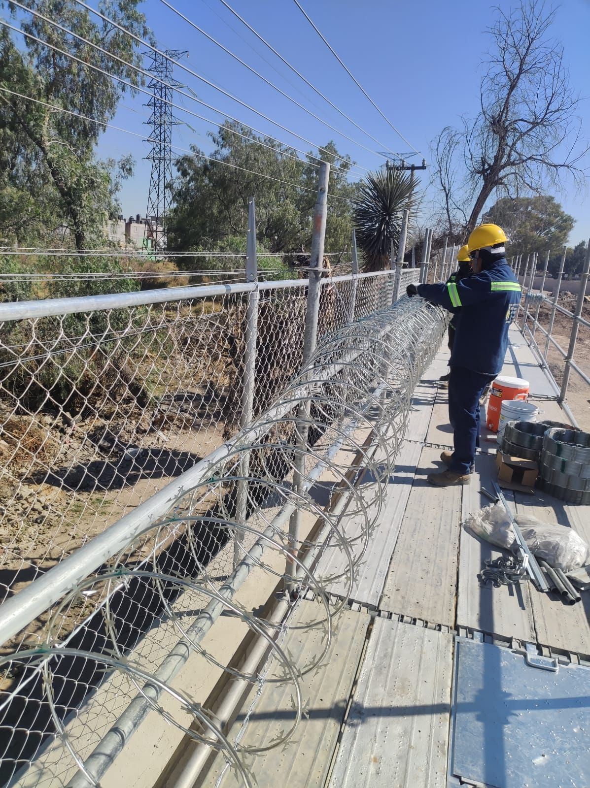 Trabajador instalando alambre de púas sobre una cerca de alambre junto a un canal. Día soleado.