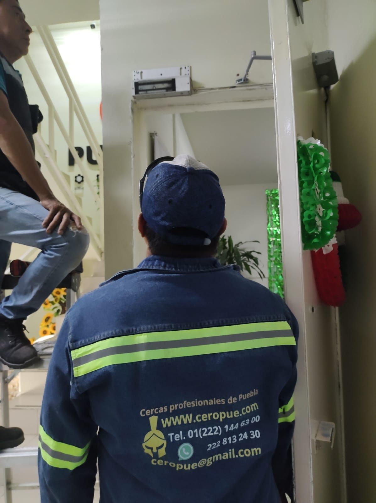 Dos hombres en un pasillo; uno de ellos con uniforme azul, mirando hacia arriba. Se ven escaleras y adornos.