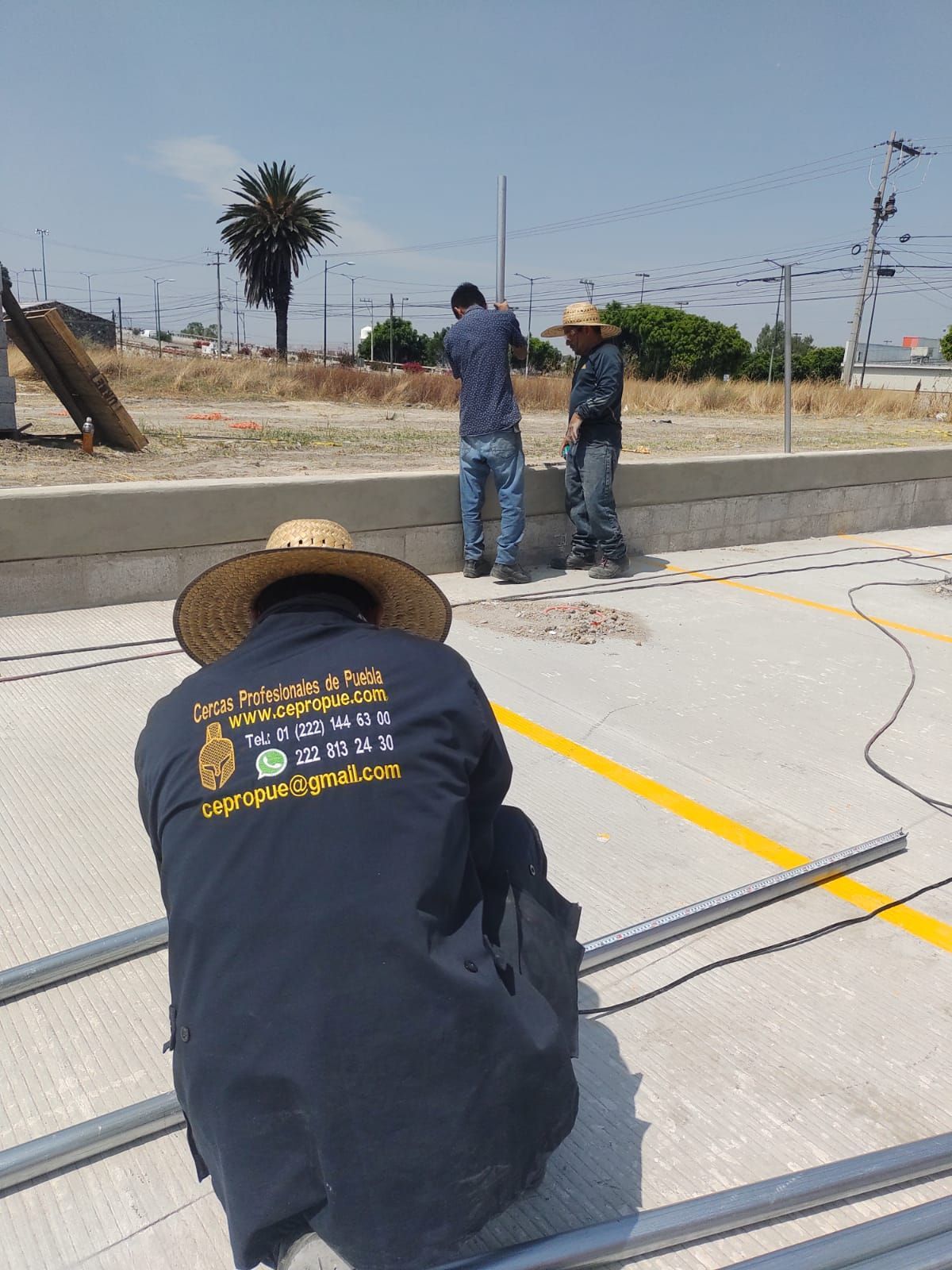 Tres trabajadores instalan estructuras metálicas sobre una superficie de hormigón. Un trabajador lleva sombrero y mira hacia abajo; dos trabajan cerca de una pared.