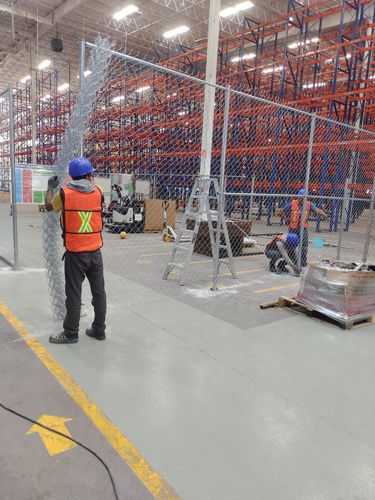 Trabajadores instalando una cerca de alambre en un almacén con estanterías altas. Un trabajador sujeta la cerca y otro se sube a una escalera.