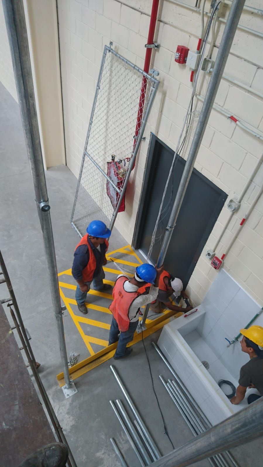 Obreros de la construcción en una plataforma cerca de una puerta. Dos llevan cascos azules y chalecos naranjas. Hay otros cerca.