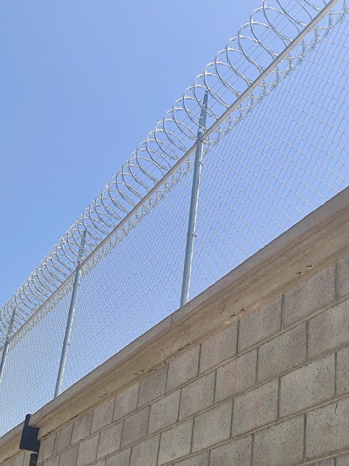 Valla de alambre de eslabones de cadena con alambre de púas sobre una pared de ladrillos contra un cielo azul.