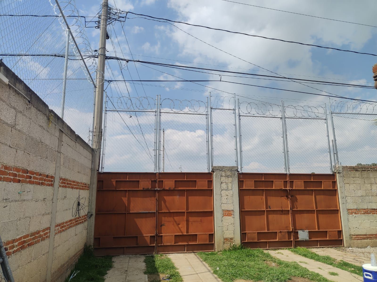 Puertas de metal oxidadas, paredes de color canela y una cerca de alambre, bajo un cielo azul con cables eléctricos encima.