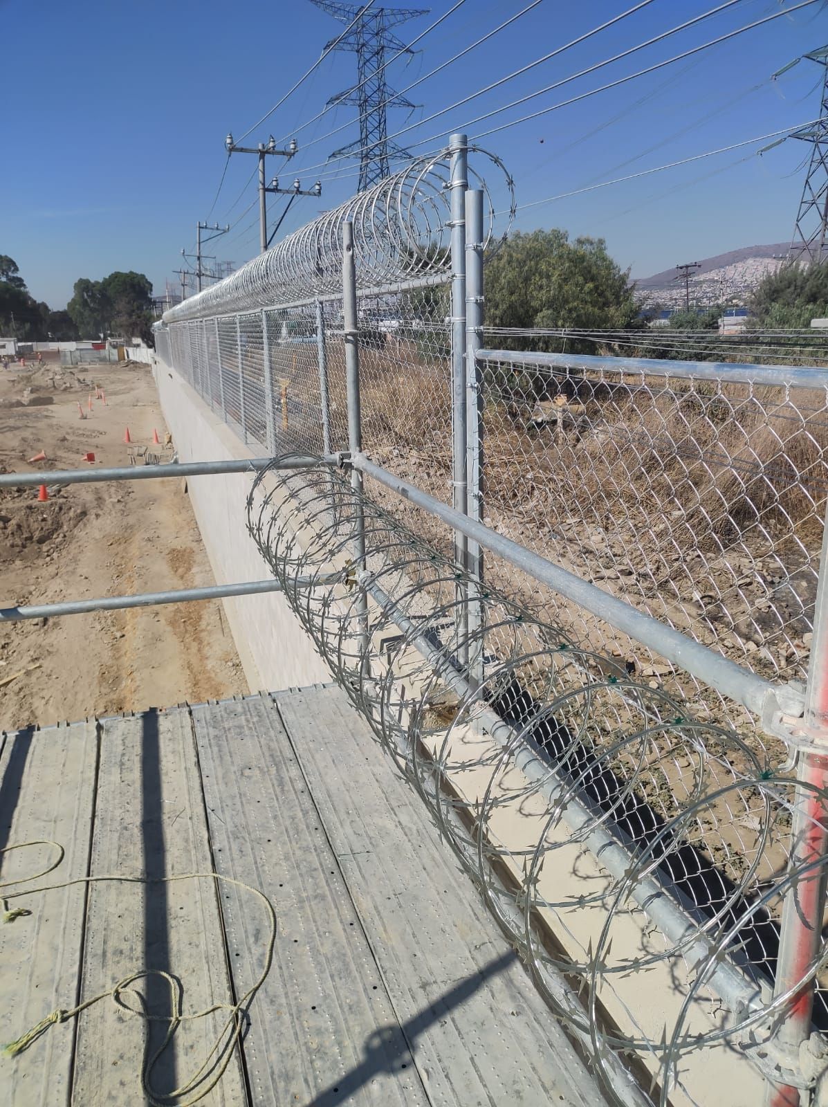 Valla de alambre de eslabones con alambre de púas sobre una barrera de hormigón, al lado de un camino de tierra y un sitio de construcción.