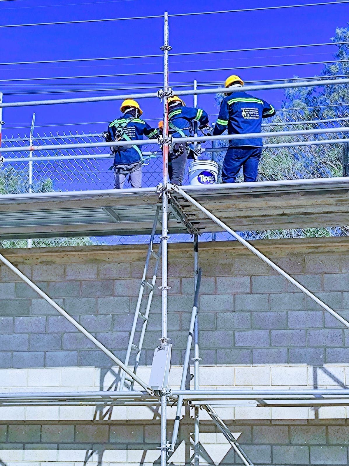 Tres trabajadores en un andamio cerca de una pared de ladrillos, con cascos y chalecos de seguridad.