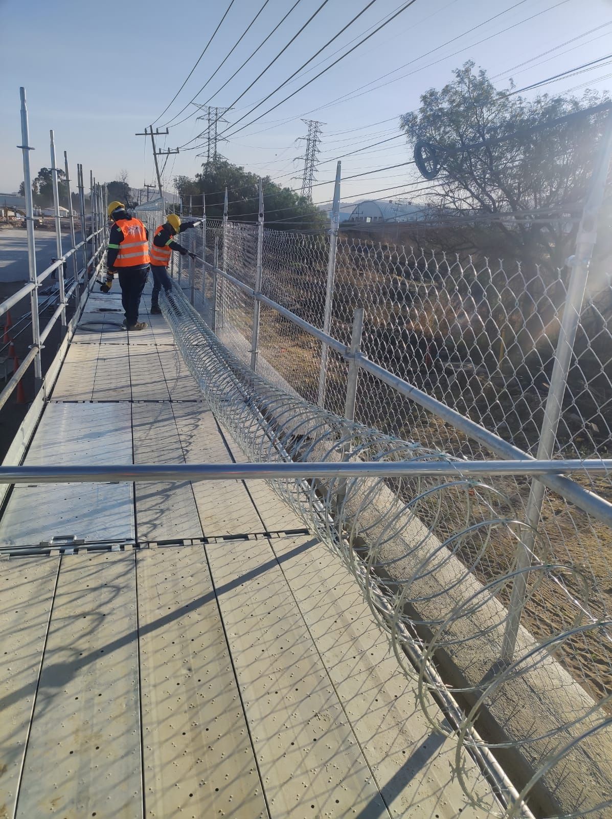 Obreros de la construcción instalando una valla de alambre a lo largo de una pasarela, día soleado.
