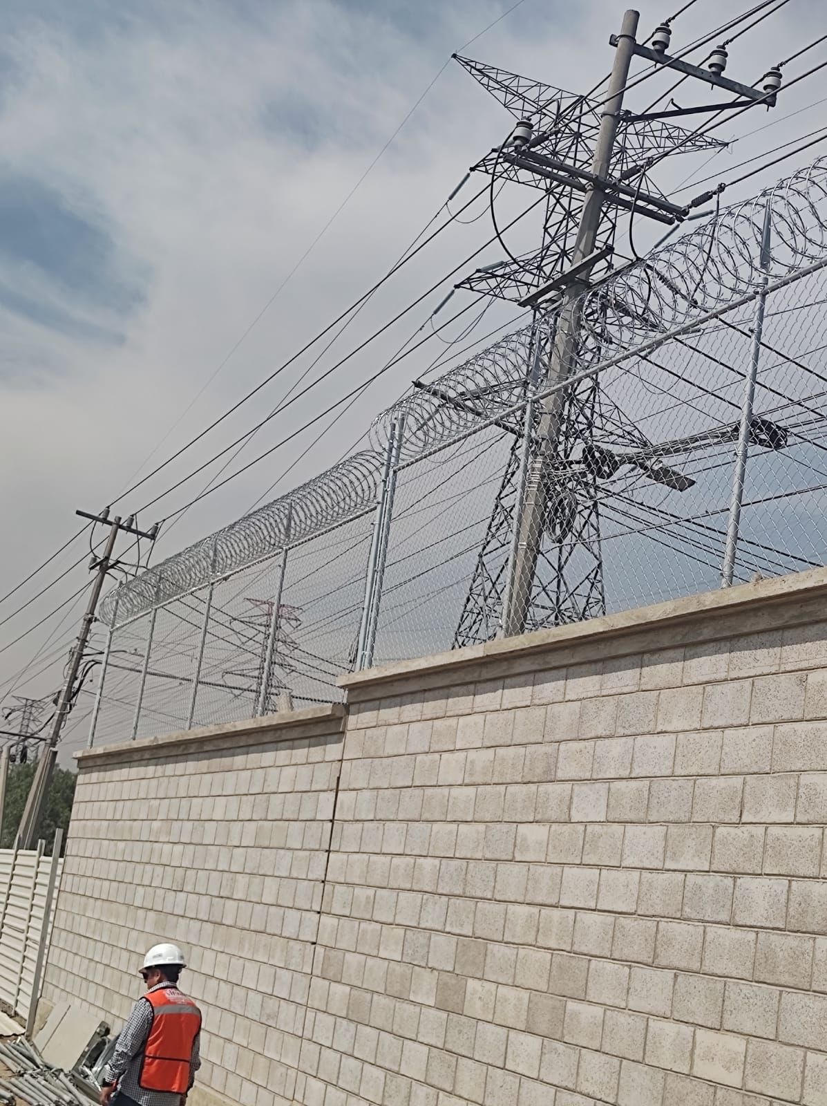Hombre con uniforme de trabajo junto a una red eléctrica de alta tensión, tras un muro de ladrillos y alambre de púas. Cielo nublado.