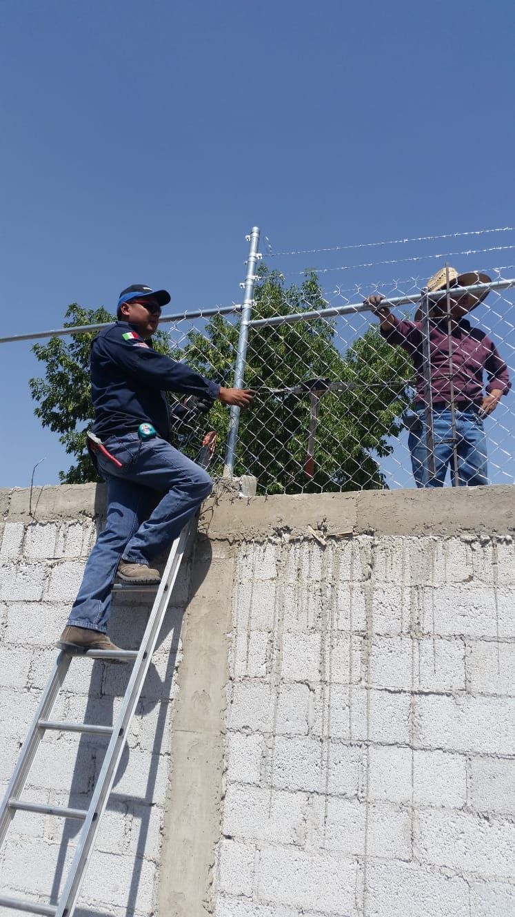 Dos hombres instalando una valla de alambre en una pared de ladrillos; uno en una escalera y el otro de pie.