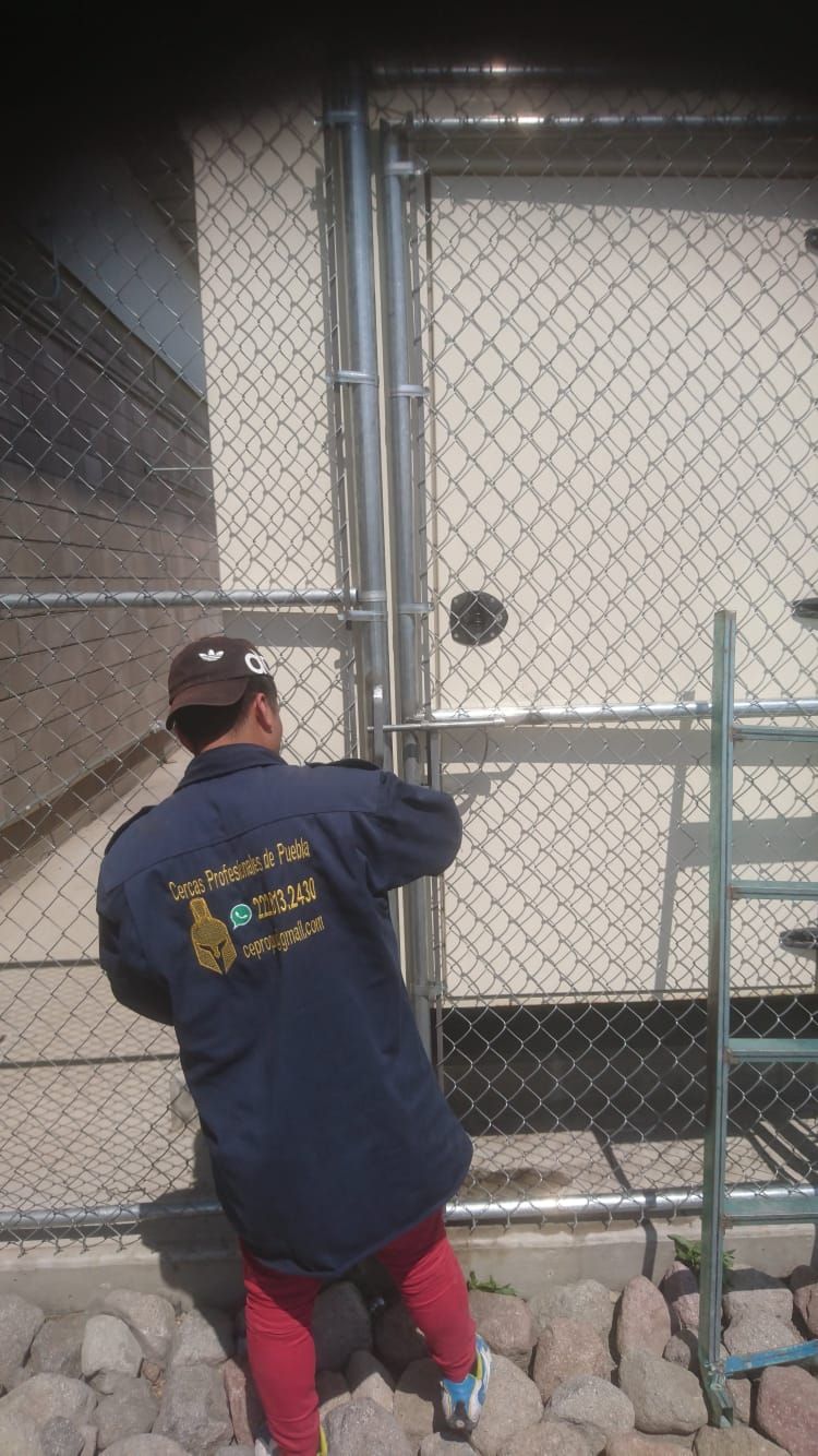 Persona con abrigo azul con logo, trabajando en una valla de alambre, al lado de una pared blanca, con una escalera.