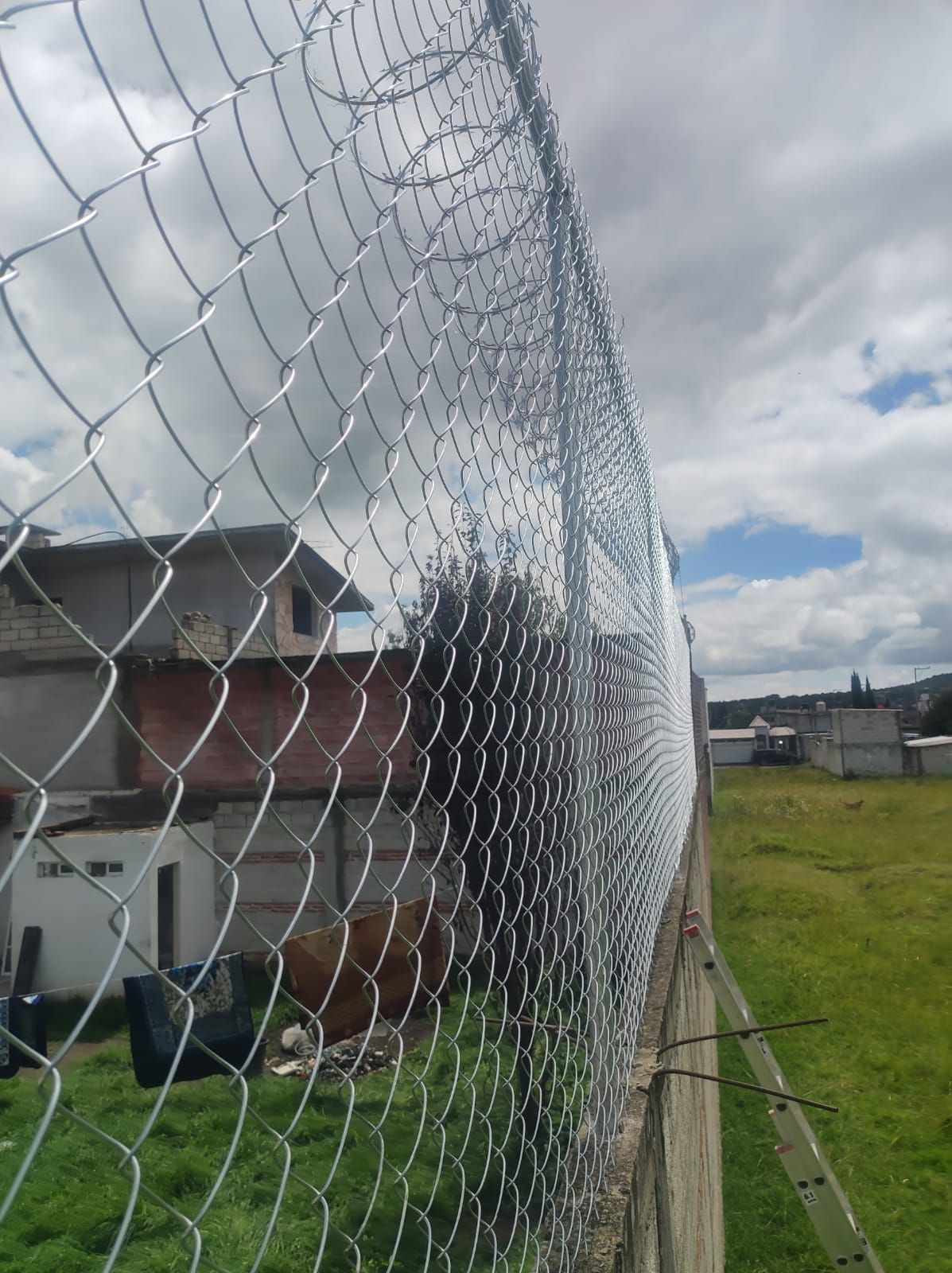 Una valla de alambre de espino, con alambre de púas encima, al lado de un campo de hierba y edificios bajo un cielo nublado.