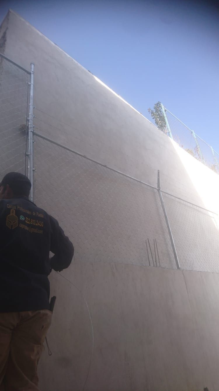 Persona con camisa negra junto a un alto muro de hormigón con una valla de alambre de púas en la parte superior.
