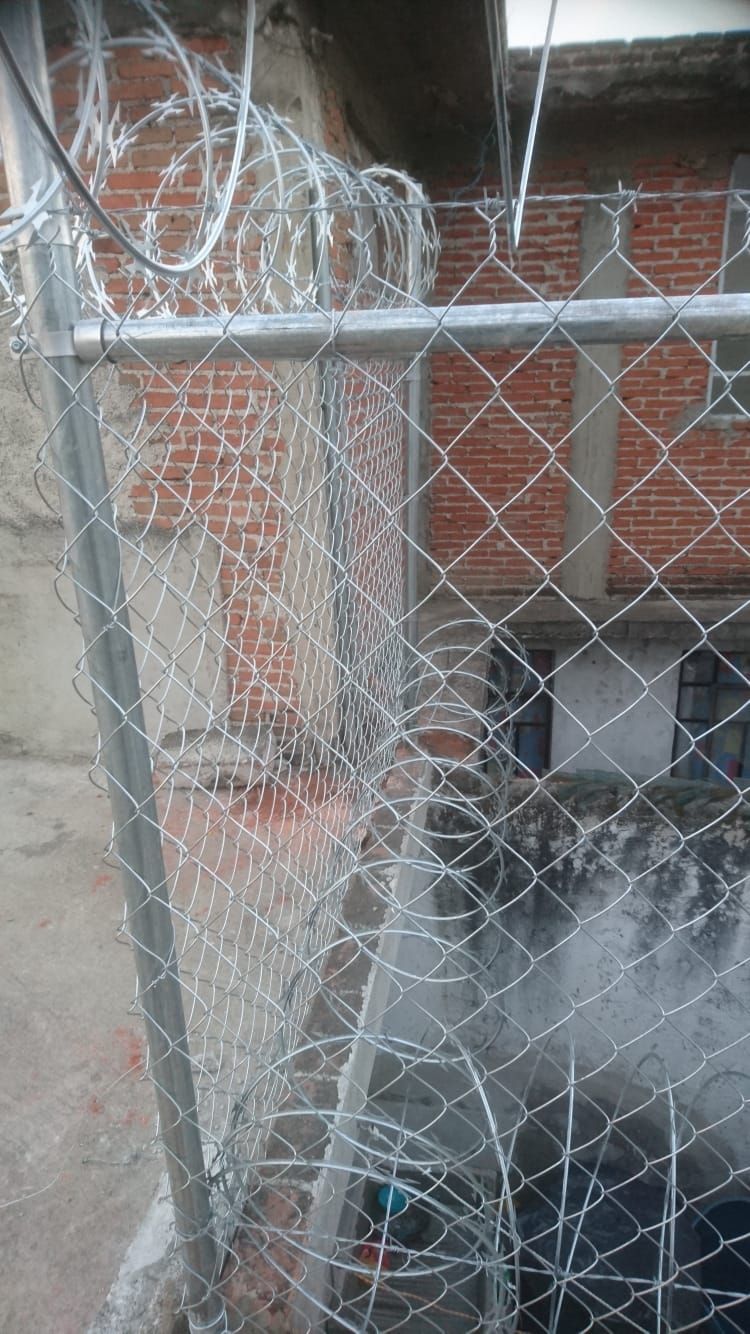 Valla de alambre de cadena rematada con alambre de púas enrollado frente a un edificio de ladrillo.