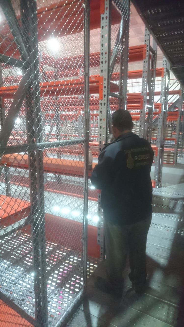 Hombre en un almacén inspeccionando estanterías metálicas con red de seguridad. Las filas de estanterías son naranjas y metálicas.