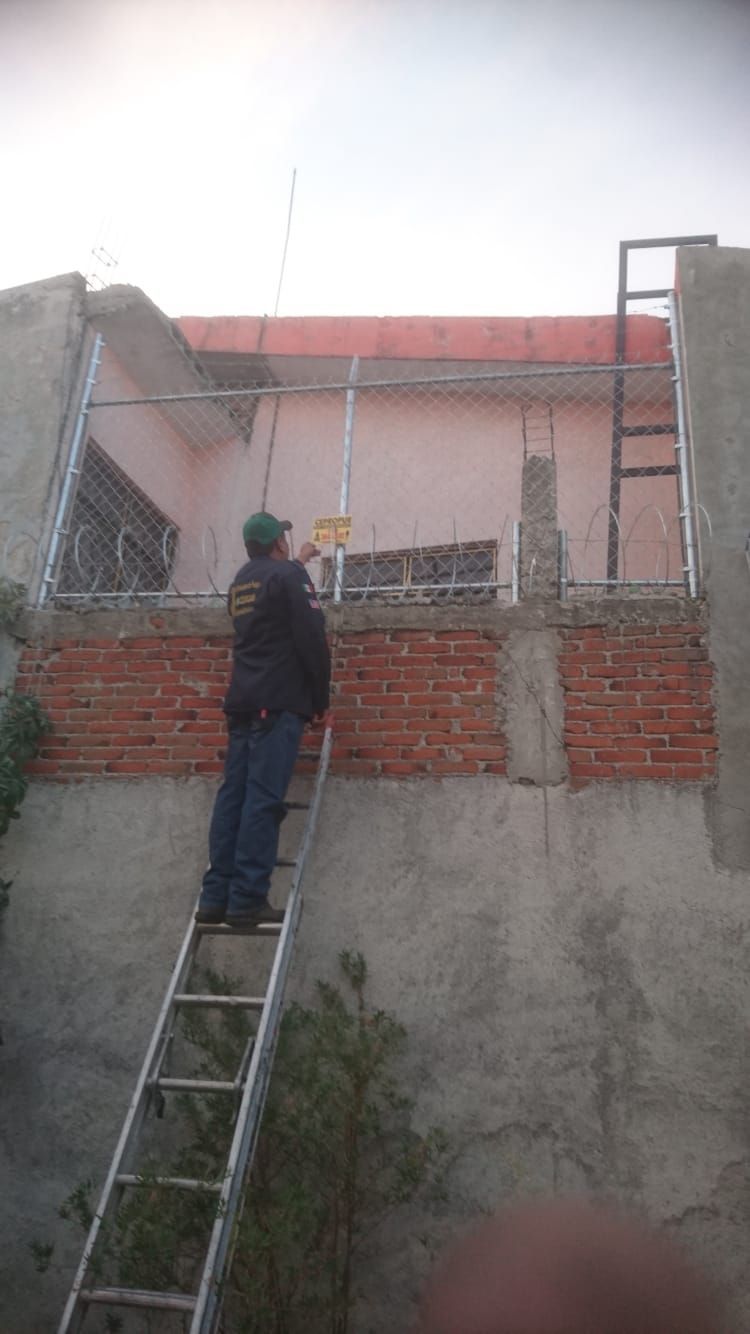Hombre en una escalera trabajando en el marco de metal de la pared exterior de un edificio, con ladrillos rojos visibles.