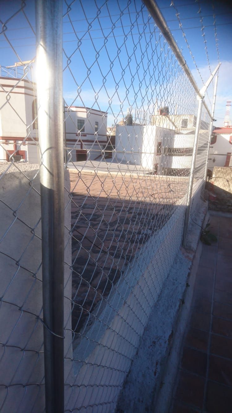 Valla de alambre en primer plano; edificios y cielo visibles al fondo.