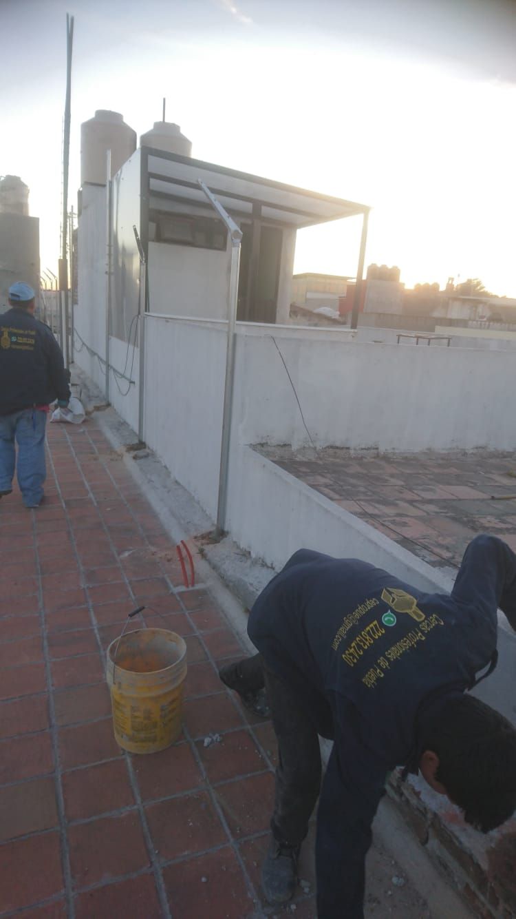 Dos hombres trabajan en una azotea con un tanque de agua y un pequeño edificio. Uno se agacha.