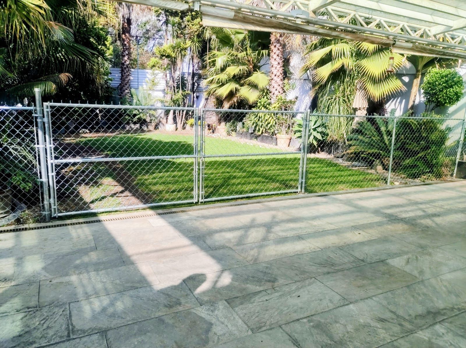 Una cerca de alambre encierra un césped verde, con árboles y un patio de hormigón en primer plano.