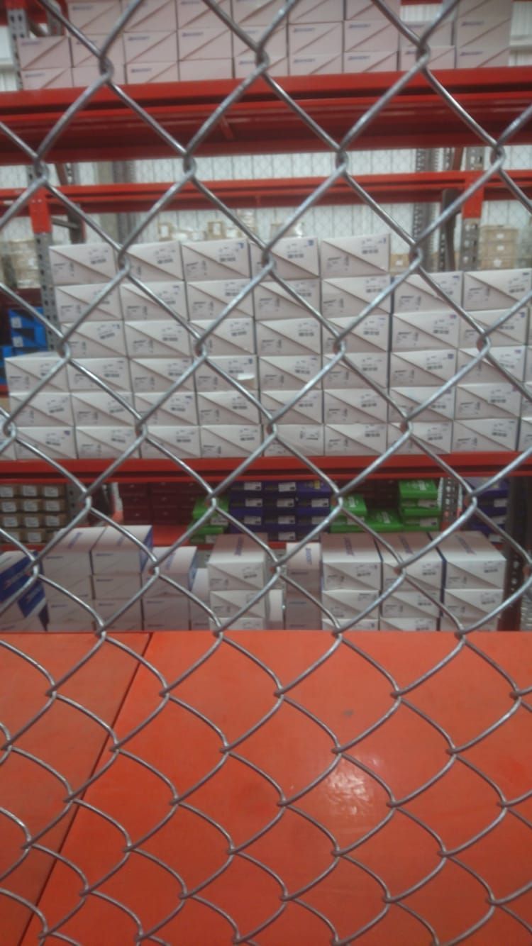 Cajas apiladas en estantes rojos detrás de una cerca de alambre, dentro de un almacén.