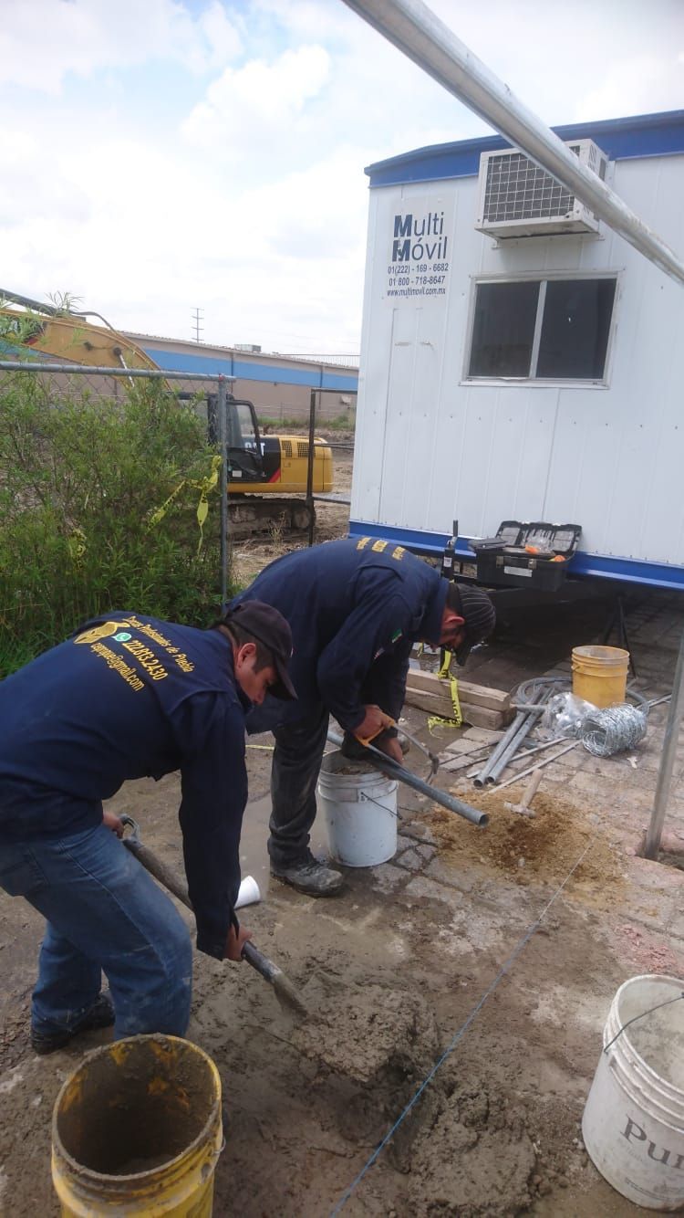 Dos trabajadores de la construcción mezclan cemento al aire libre cerca de un remolque y una pequeña excavadora.