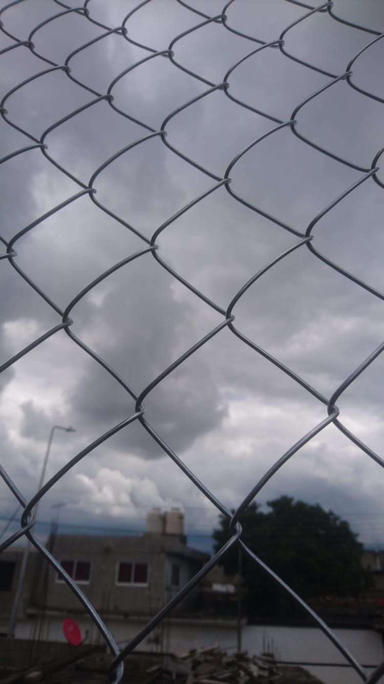 Valla de alambre contra un cielo nublado con un edificio y un árbol al fondo.