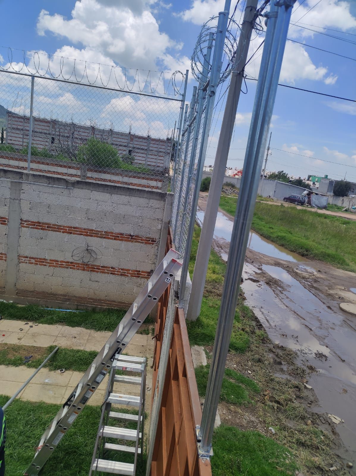 Escalera apoyada en una valla con postes metálicos nuevos y cables bajo un cielo nublado.