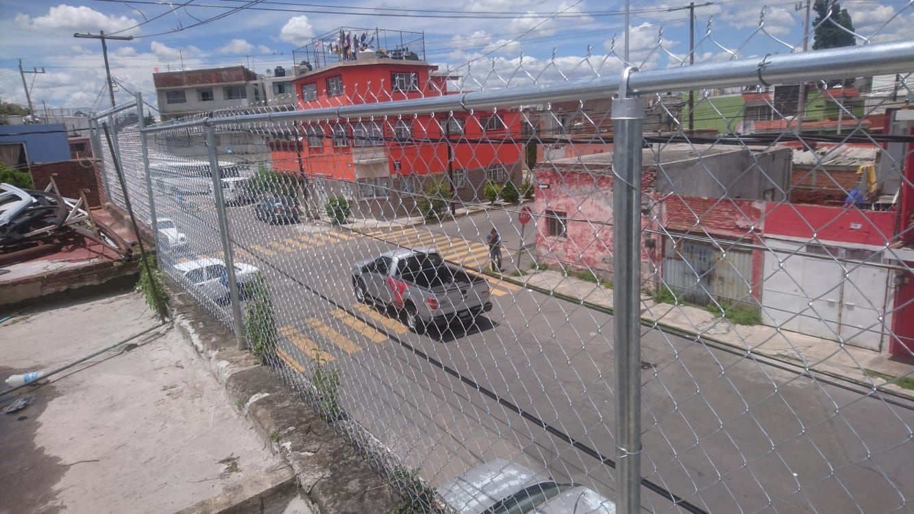 Escena callejera con una valla de alambre en primer plano, edificios al fondo y un camión circulando por la carretera.
