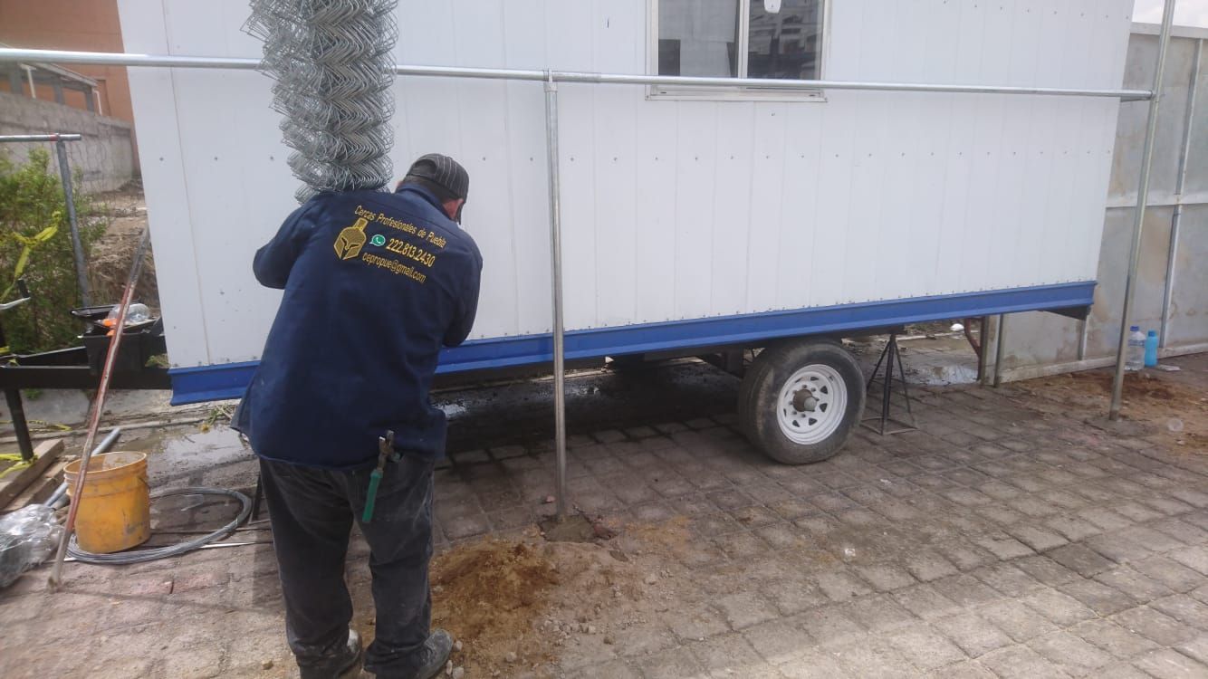 Hombre trabajando cerca de un remolque blanco con detalles azules, sobre una superficie de hormigón, al aire libre.