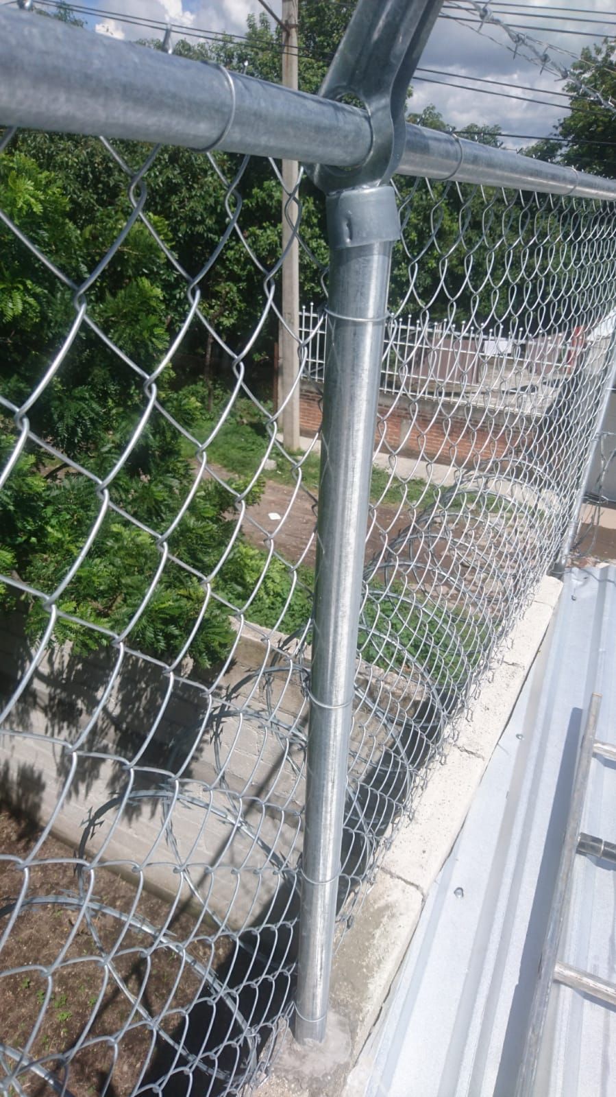 Valla de alambre con poste de metal en un tejado, con árboles verdes al fondo.