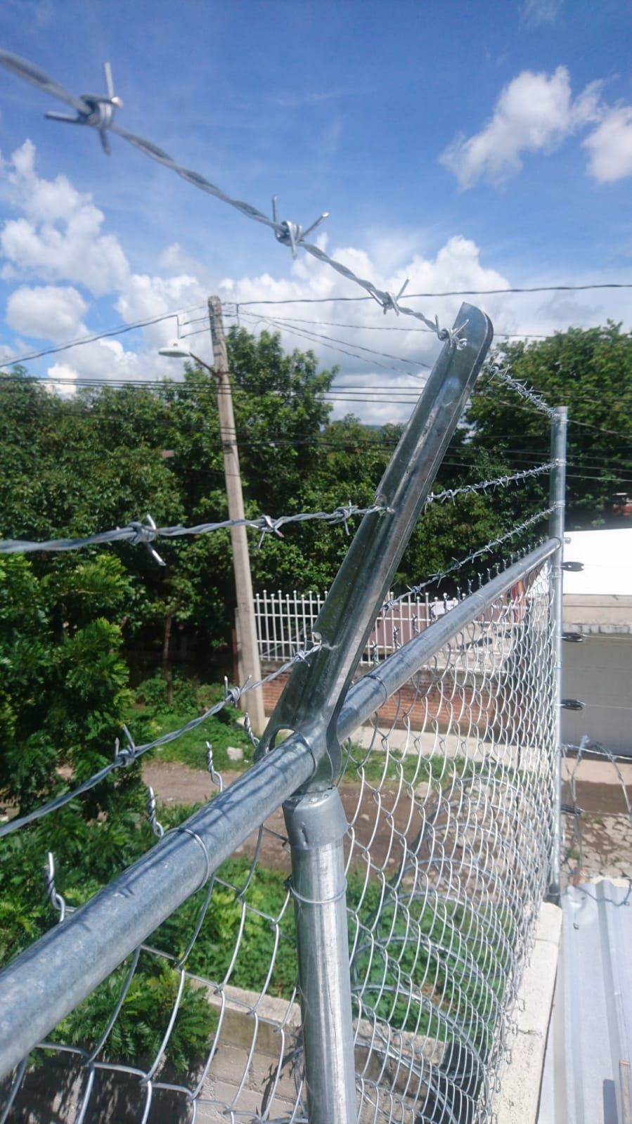 Valla de alambre de cadena rematada con alambre de púas contra un cielo nublado y árboles verdes.