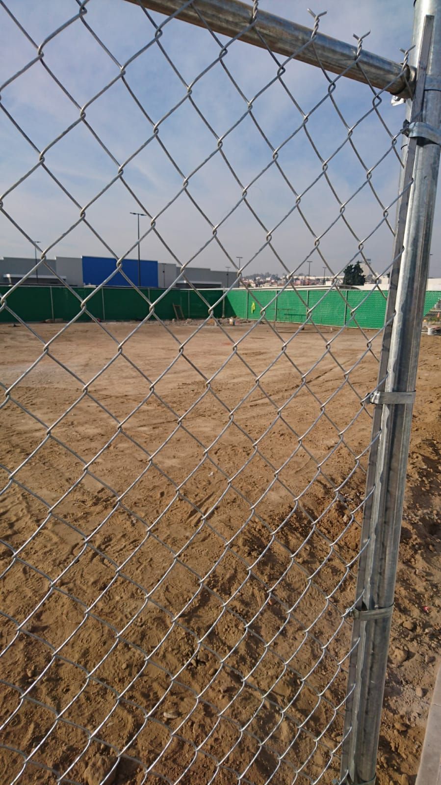 Cerca de alambre con vista a un lote de tierra, un sitio de construcción y edificios azules y verdes más allá en un día soleado.