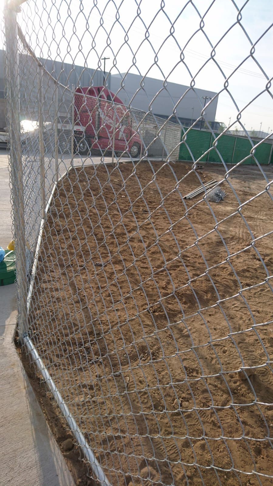 Una valla de alambre rodea una zona arenosa; al fondo se ven un camión rojo y edificios industriales.