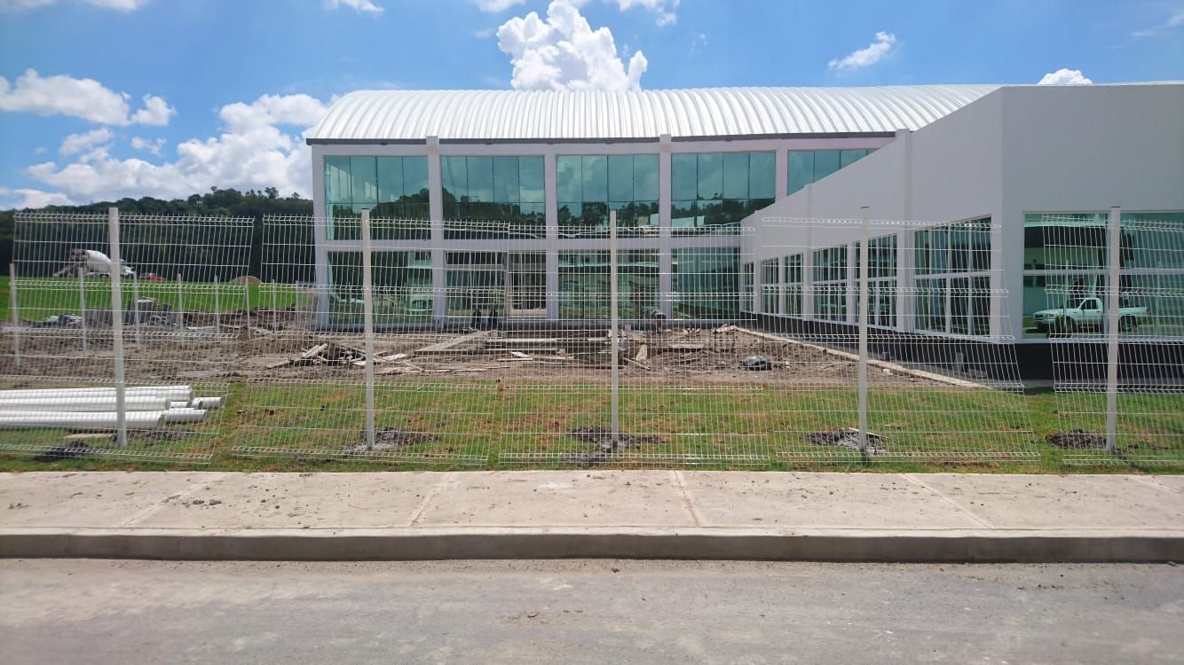 Un edificio moderno y blanco con ventanas de cristal y una valla metálica al frente. Cielo azul nublado.