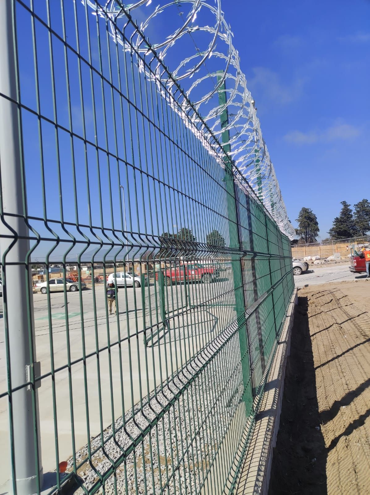 Valla de seguridad verde con alambre de púas bajo un cielo azul.